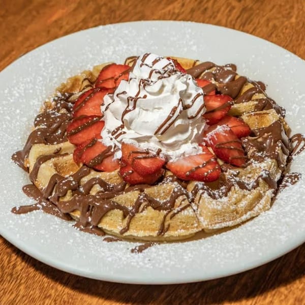 JJ Cafe waffle topped with strawberries, whipped cream, chocolate drizzle, and powdered sugar.